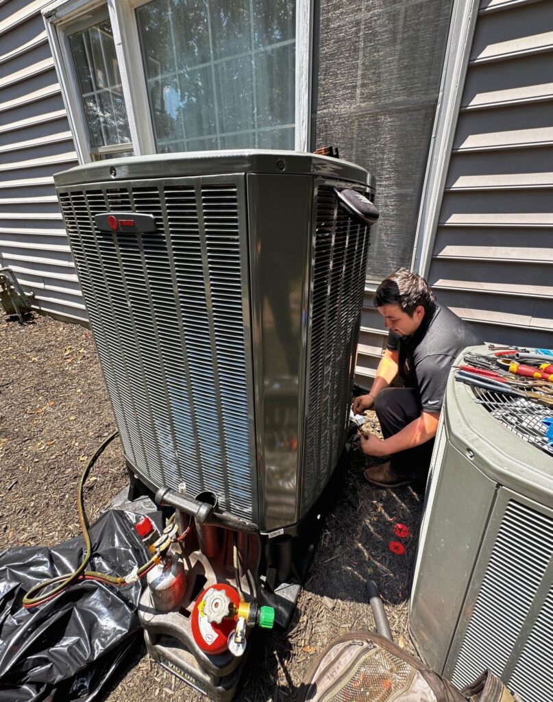A SwiftPro technician crouched at the side of an outdoor air conditioning condenser in Fairfax, Virginia, with refrigerant gauges and a tool roll on the ground beside him