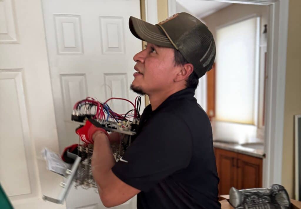 A SwiftPro heating technician carrying an electric heat strip assembly through a Northern Virginia home on a heat pump repair