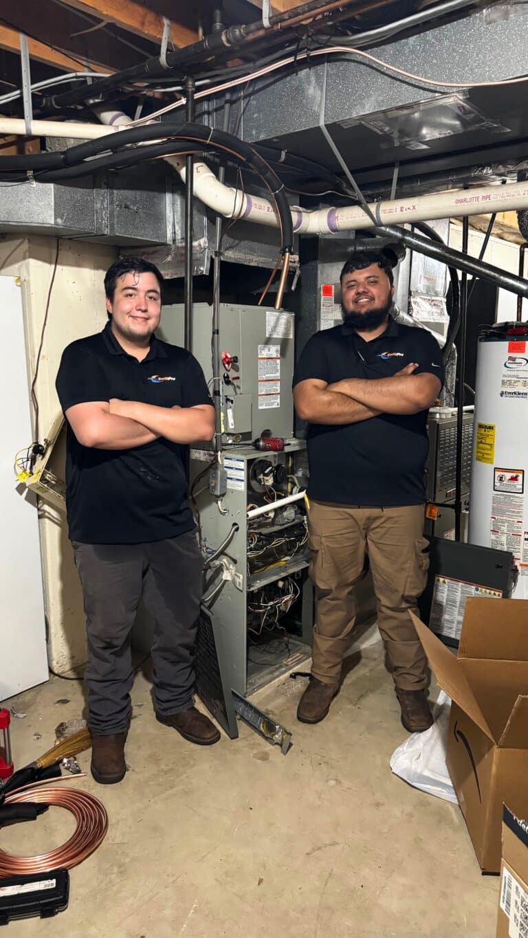 Two SwiftPro HVAC technicians in black company polos standing arms crossed in a Northern Virginia basement, beside a gas furnace with its front panel off and its wiring open, a water heater to the right