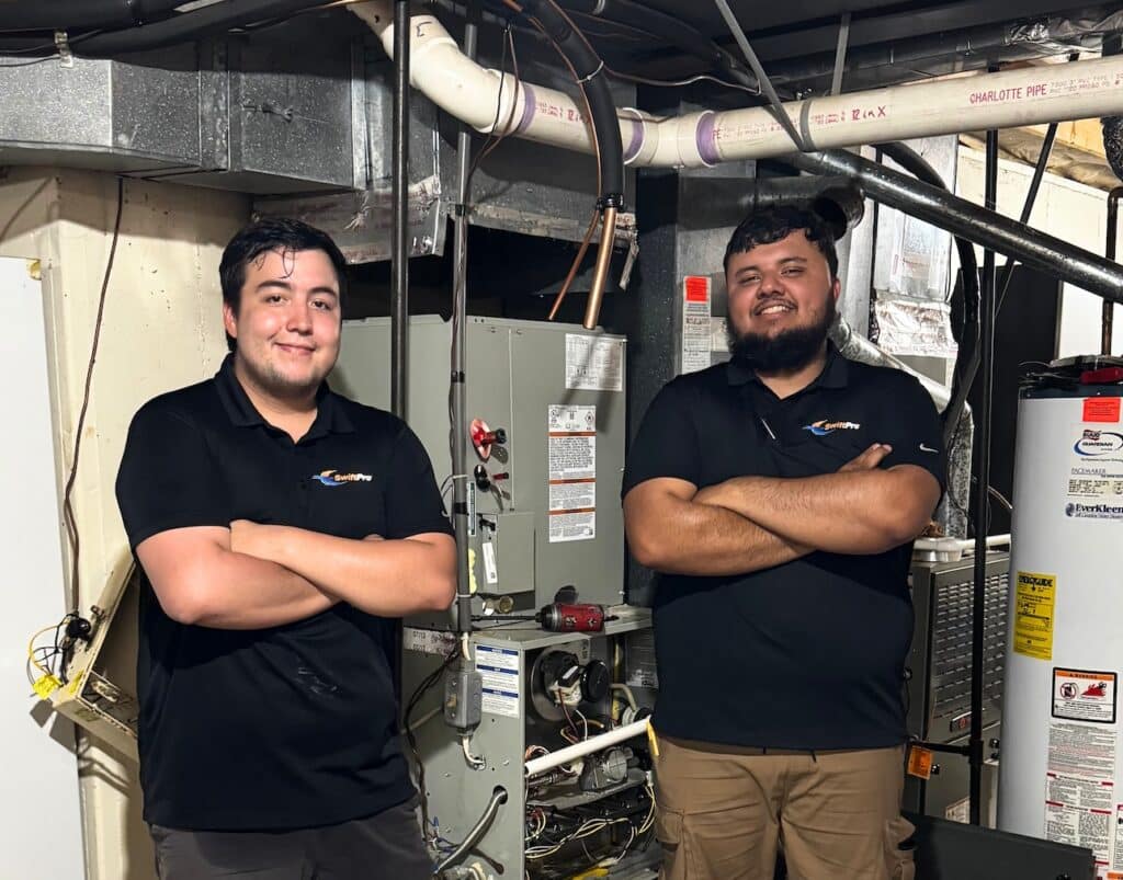 Two SwiftPro technicians standing in a residential mechanical room beside an air handler and a water heater