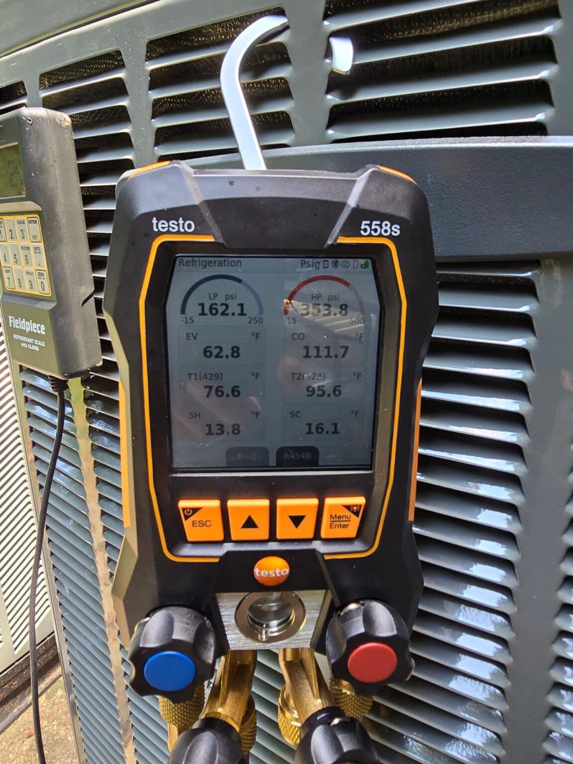 A digital refrigerant manifold hanging on an outdoor AC unit, reading suction and liquid line pressures and temperatures