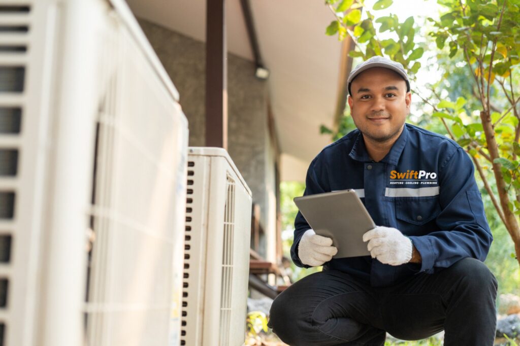 A SwiftPro technician in a branded polo kneeling beside an outdoor unit with a tablet in hand