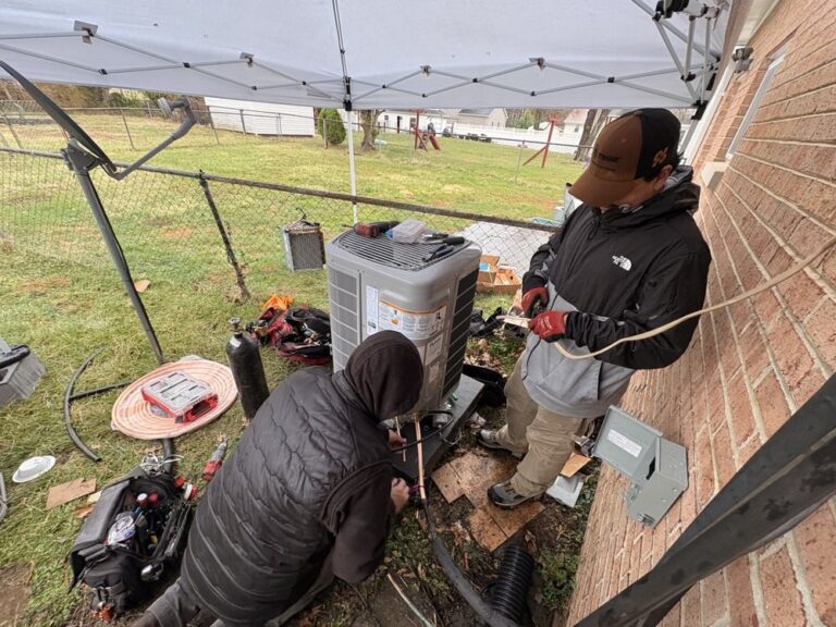 Two SwiftPro technicians working on an outdoor heat pump under a canopy in wet weather.
