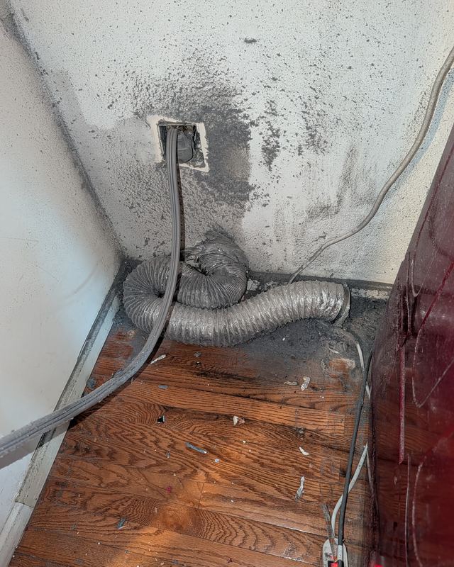 Behind a dryer in an Ashburn, Virginia laundry room: a crushed foil dryer hose and grey lint around the vent opening in the wall, before a SwiftPro dryer vent cleaning