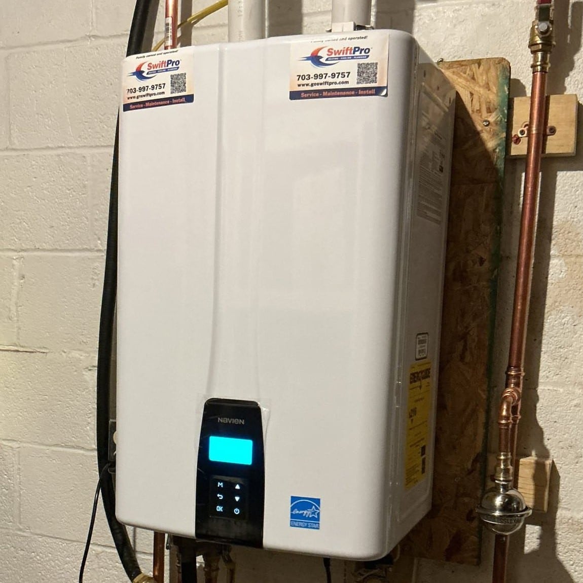 A Navien tankless water heater on a basement wall with SwiftPro service stickers on its front