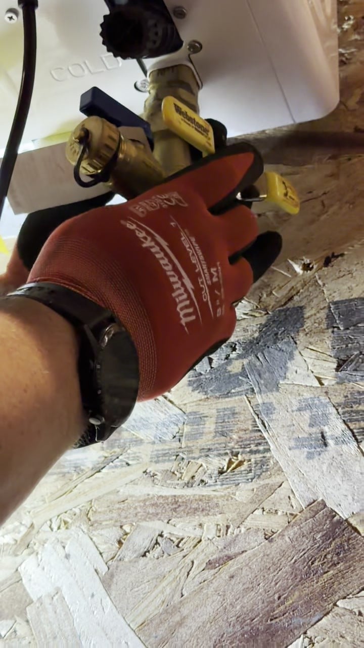 A gloved hand at the yellow handled isolation valves under a tankless water heater, the valves used to flush it