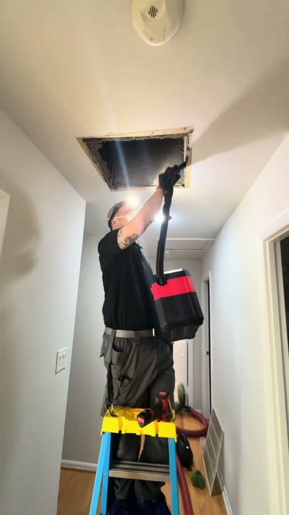 A SwiftPro technician on a ladder feeding a vacuum hose up into an opened ceiling duct in a Northern Virginia home, his headlamp lighting the opening