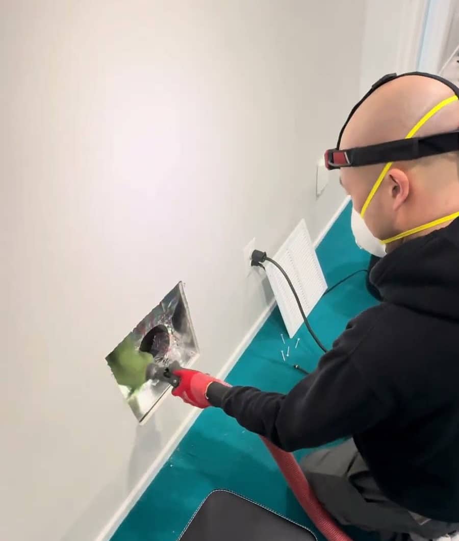 A SwiftPro technician in a headlamp and dust mask feeding a vacuum hose into an opened wall register while cleaning the air ducts in a Northern Virginia home