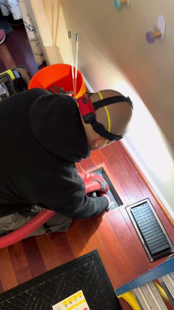 A SwiftPro technician kneeling at an open floor register in a Northern Virginia home, the vacuum hose run into the duct below
