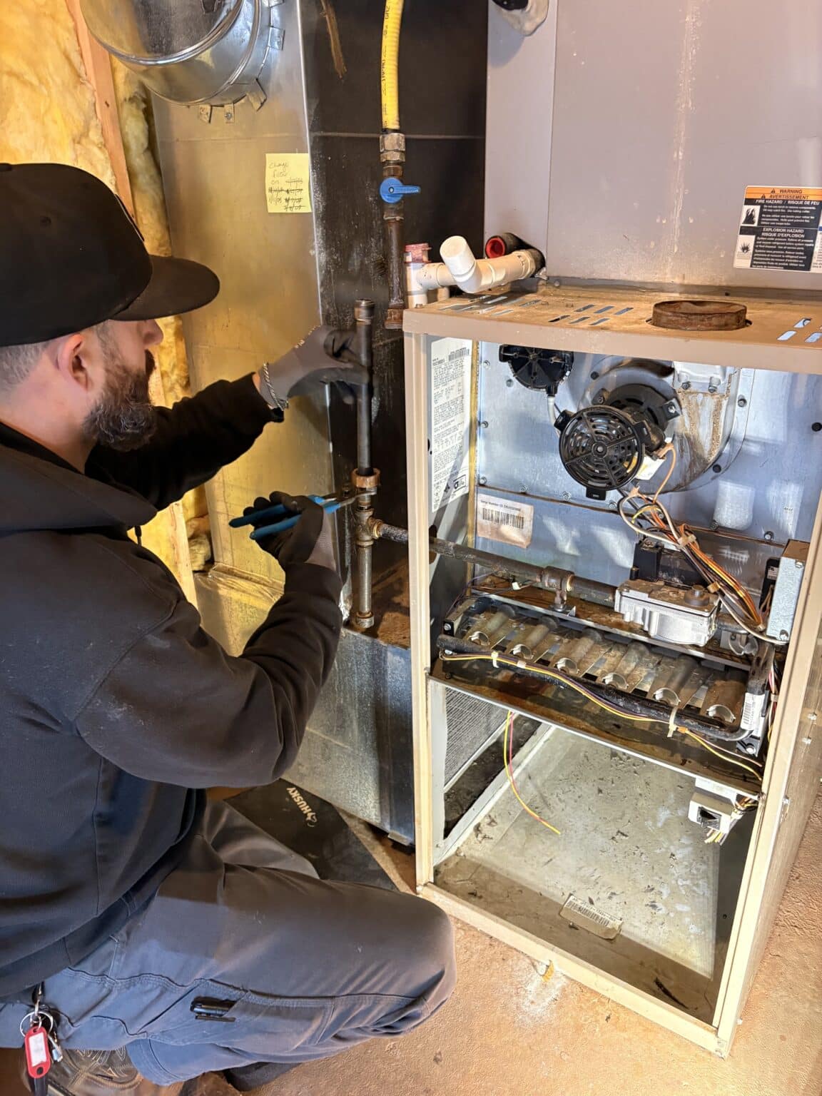 A SwiftPro technician in a black cap working on the gas piping beside an open gas furnace in a Northern Virginia home, the burners, inducer and gas valve visible
