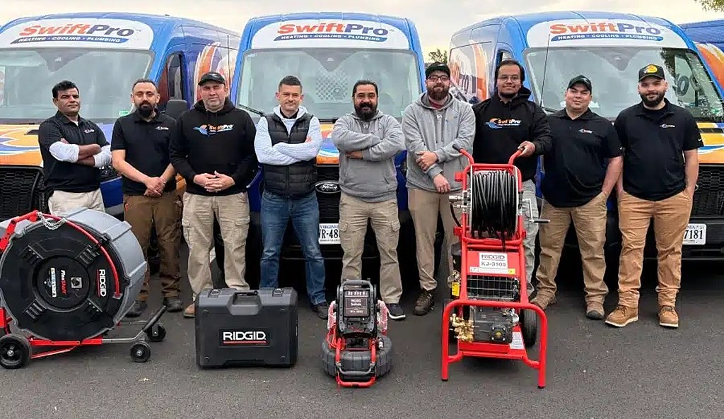 Nine of the SwiftPro crew lined up in front of the wrapped SwiftPro vans, with a drain machine and a water jetter