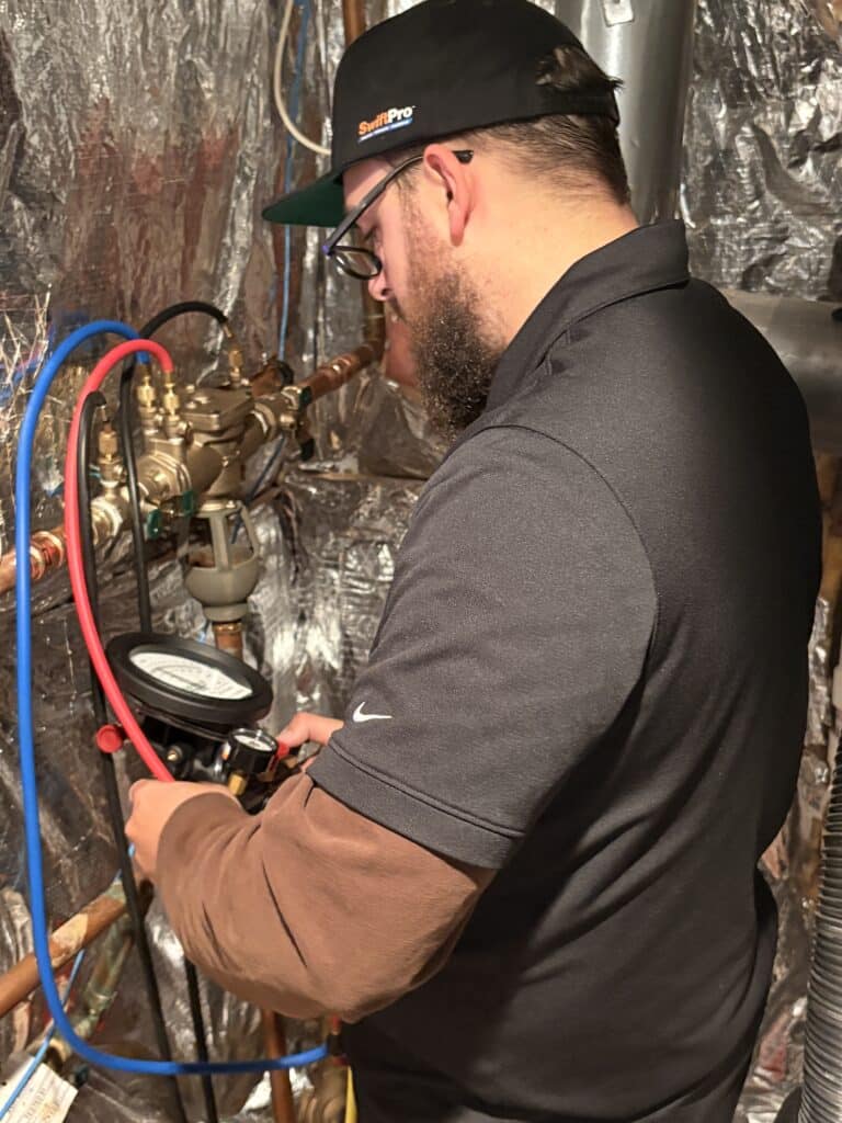 A SwiftPro plumber in a SwiftPro cap holding a backflow test kit gauge, its blue, red and black hoses connected to the test cocks of a brass RPZ backflow preventer in an Ashburn, Virginia basement
