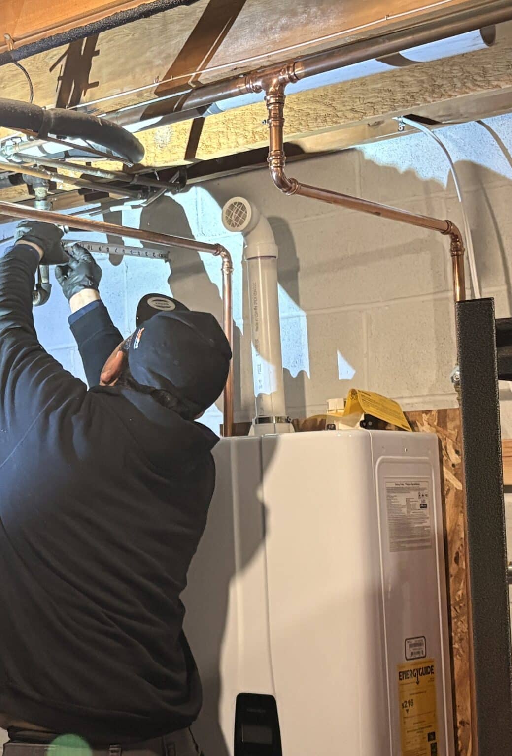 A SwiftPro plumber fitting new copper water lines overhead in a Northern Virginia basement, a new condensing tankless water heater below him with its white plastic venting on top