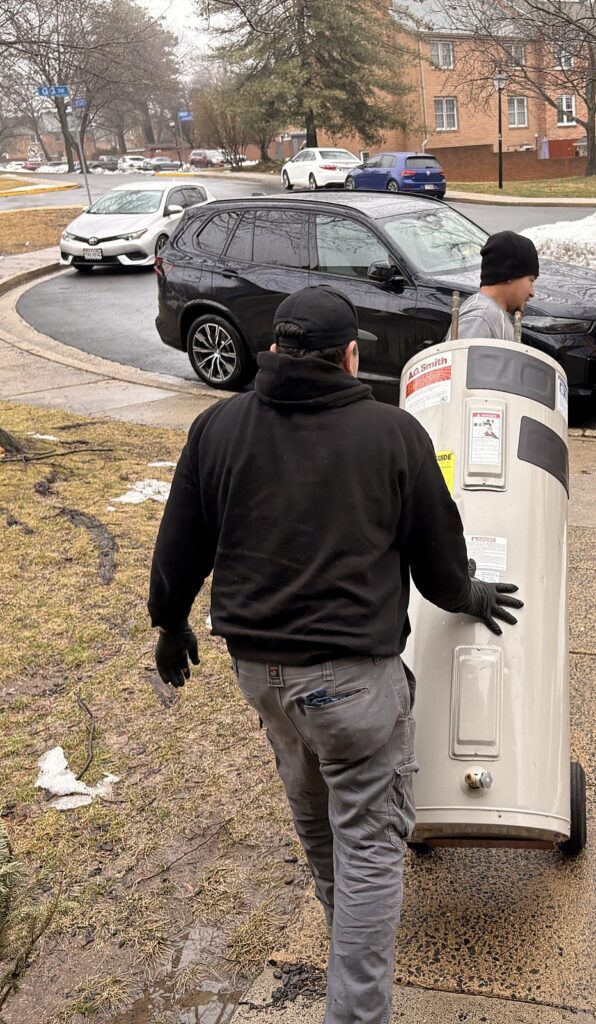 A SwiftPro technician carrying a new tank water heater up the front walk of a Northern Virginia home, the wrapped van behind