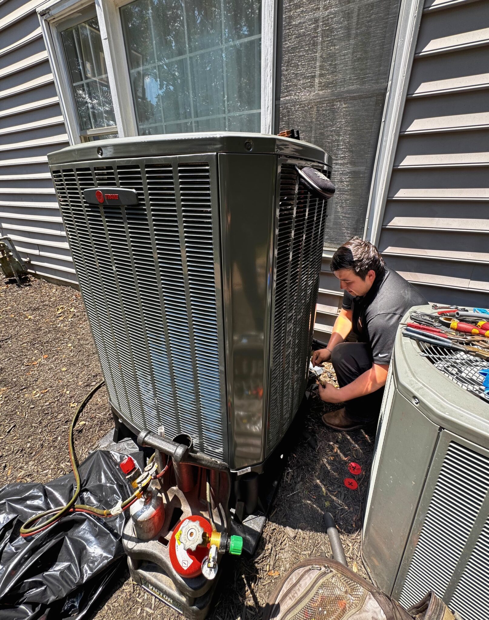 A SwiftPro technician kneeling at the base of a Trane outdoor unit beside a Northern Virginia house, refrigerant tanks, hoses and tools laid out on the ground