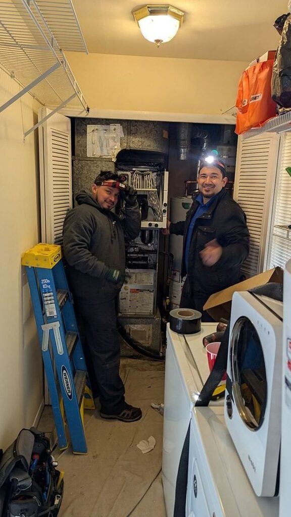 Two SwiftPro technicians at an open furnace in the utility closet of a Northern Virginia home, the water heater behind them