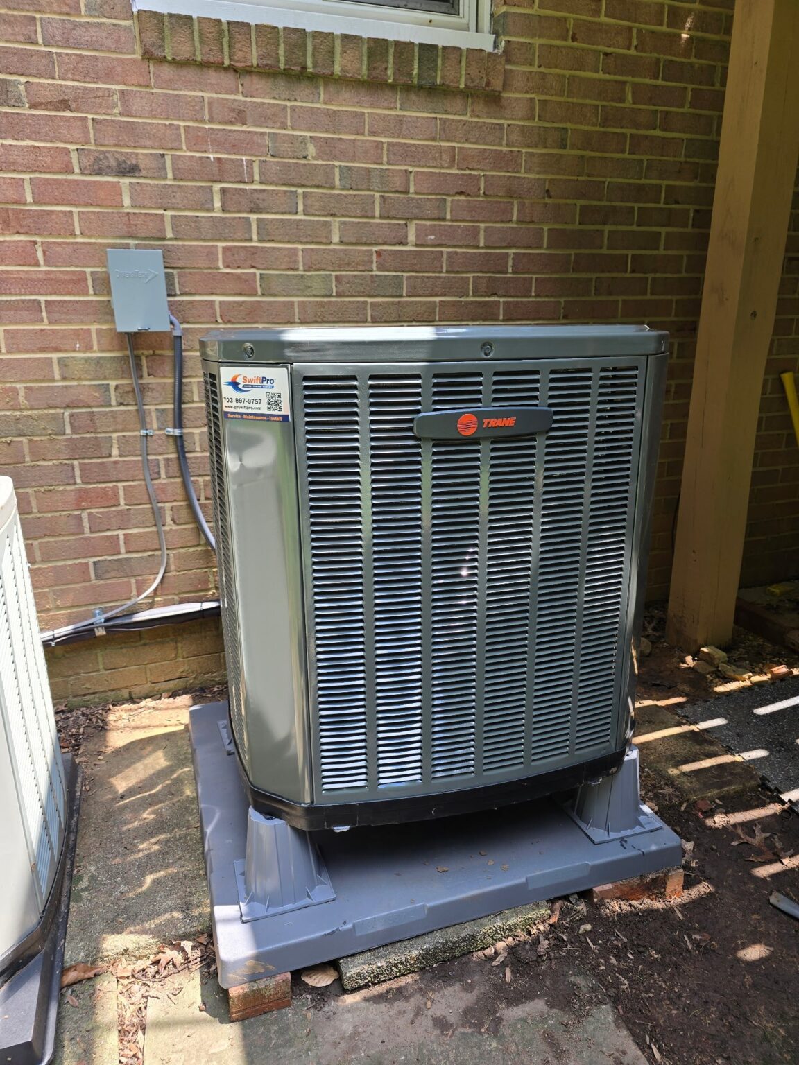 A new Trane outdoor unit on a raised pad against a brick wall in Northern Virginia, with a SwiftPro service sticker on its corner panel