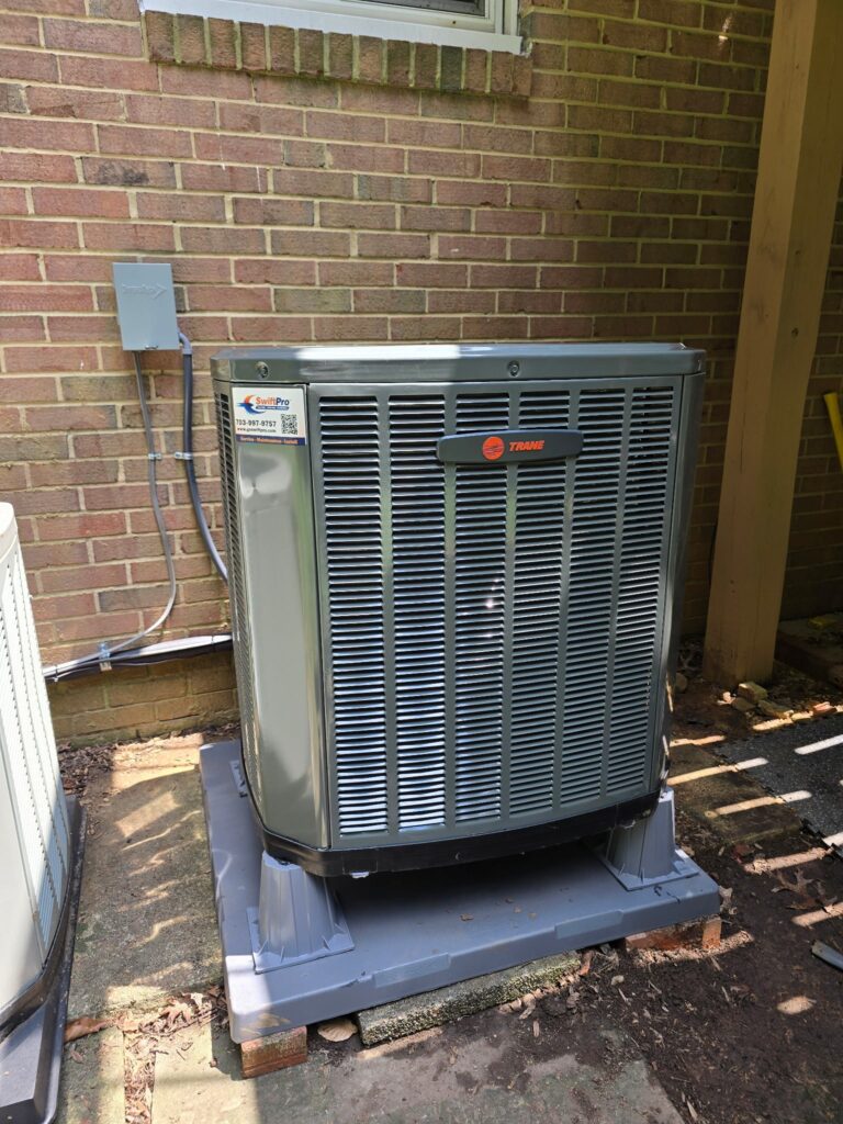 A new Trane outdoor unit on a raised pad against a brick wall in Northern Virginia, with a SwiftPro service sticker on its corner panel