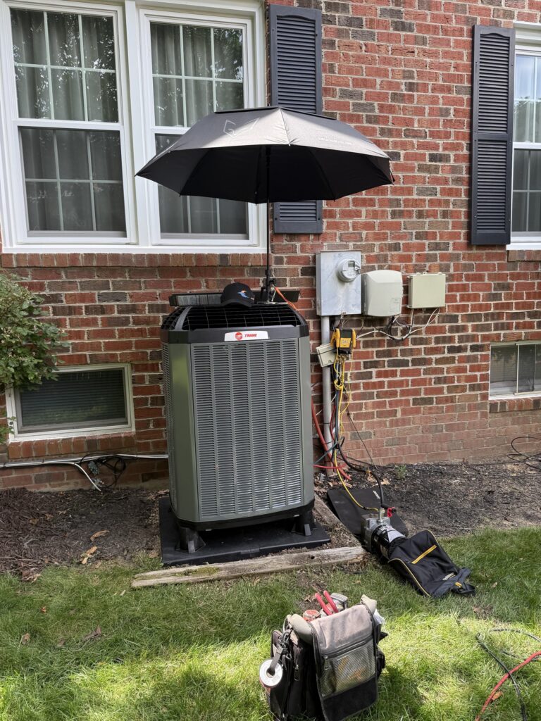 A new Trane outdoor unit being installed beside a brick house in Northern Virginia, refrigerant gauges hooked up, an umbrella shading the work and tool bags on the grass