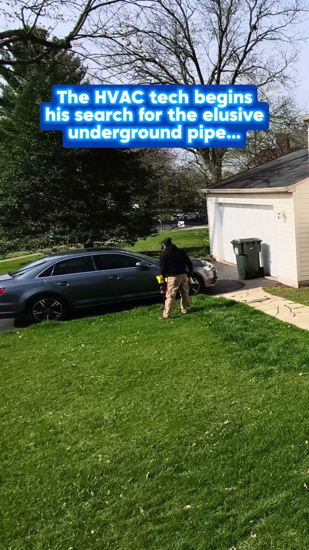 A tech sweeps a pipe-locating wand across a front lawn beside a parked car and a detached garage, hunting for a buried pipe.