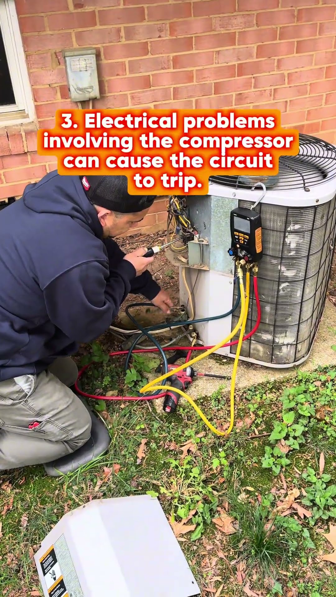 A tech in a navy hoodie and beanie hooks digital manifold gauges to an outdoor AC condenser beside a brick house with the service panel off; close-up of hands on the refrigerant lines.