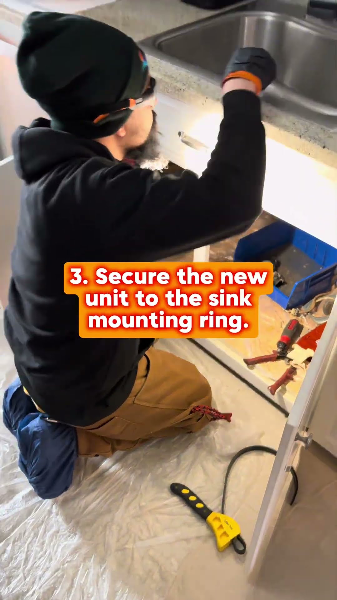 Garbage disposal replacement in a kitchen: a tech in a black hoodie and beanie kneels under the sink on a drop cloth, secures the new unit to the mounting ring, and runs water to check for leaks.