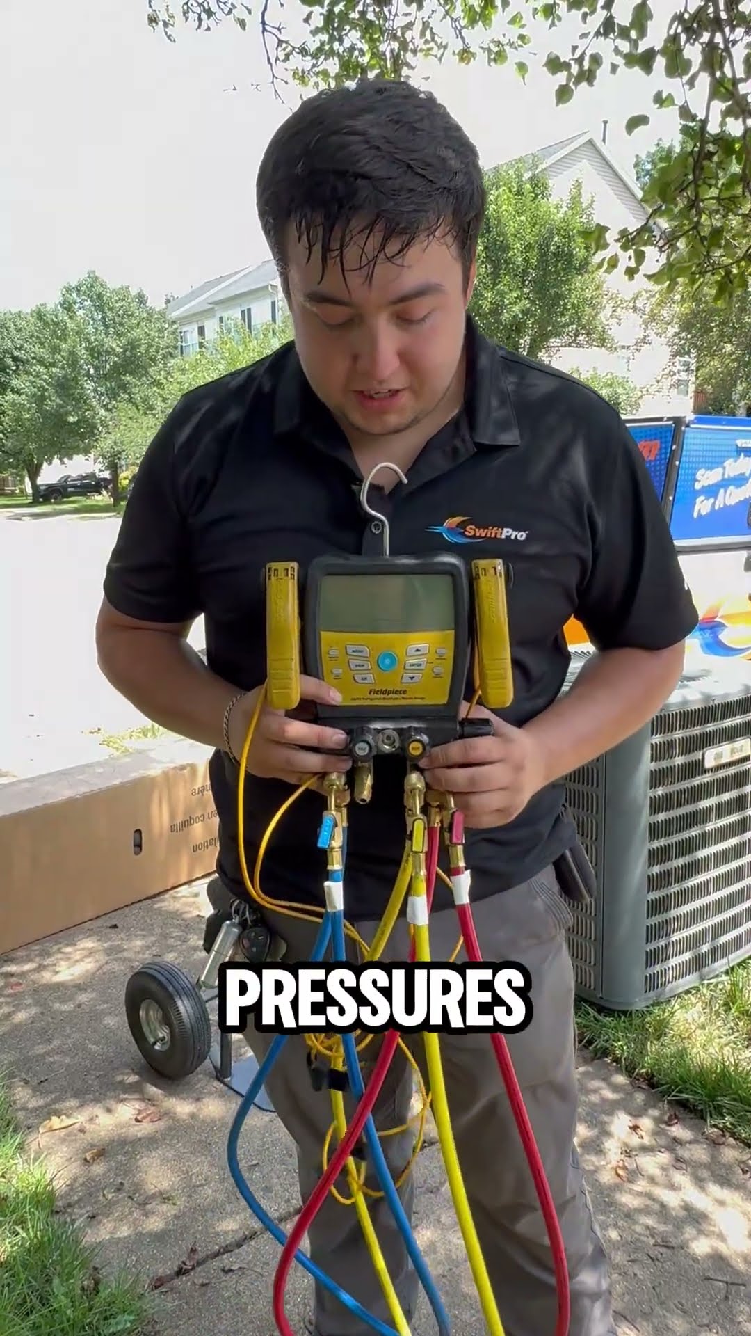 A young tech in a black SwiftPro polo stands beside an outdoor AC unit on a residential sidewalk holding a digital refrigerant manifold gauge with red, blue and yellow hoses, explaining how gauges rea