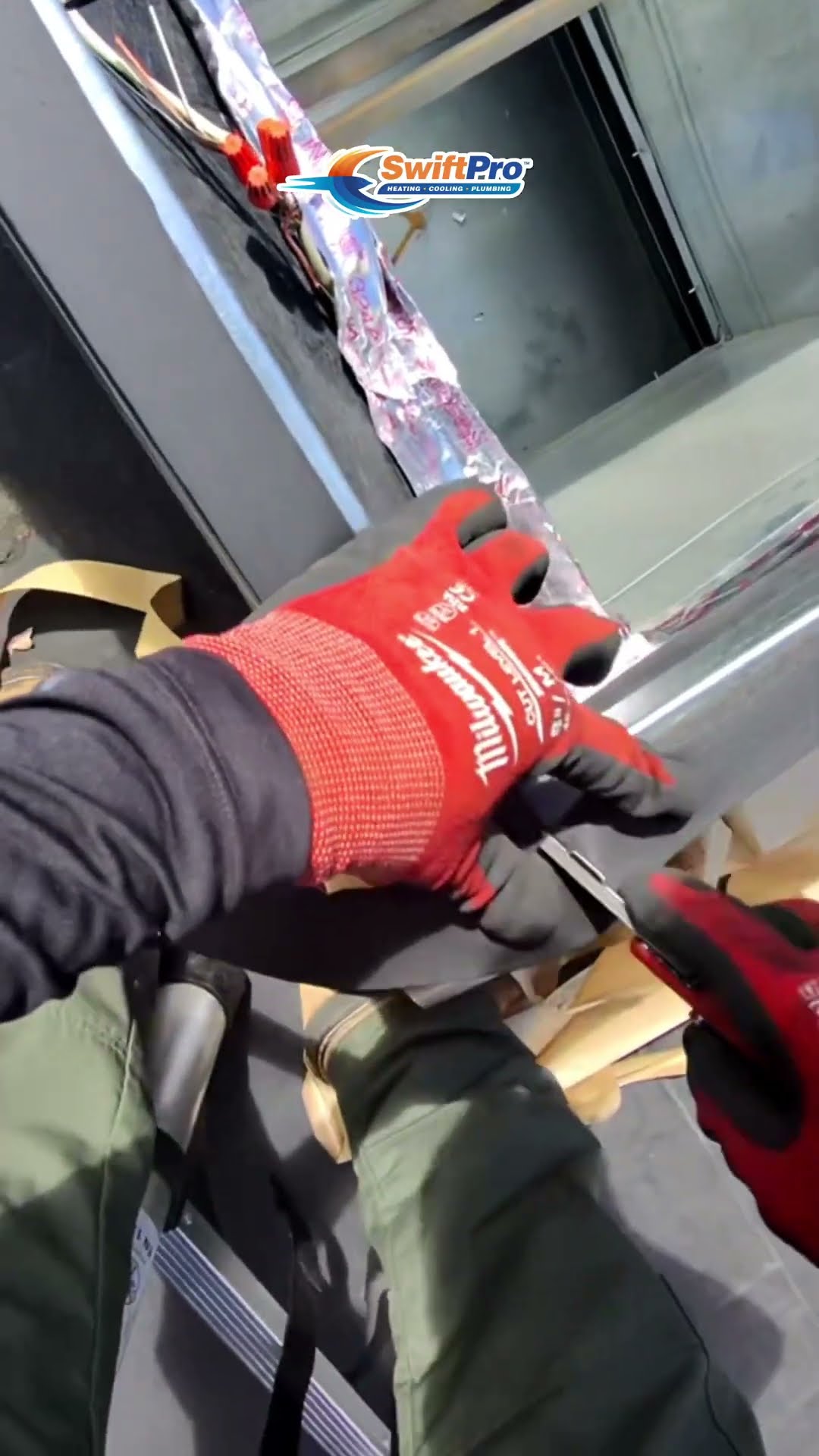 First-person view on a roof: hands in Milwaukee gloves seal the foil-lined duct connection on a rooftop unit's curb and strip and join control wiring, with a DeWalt tool nearby.