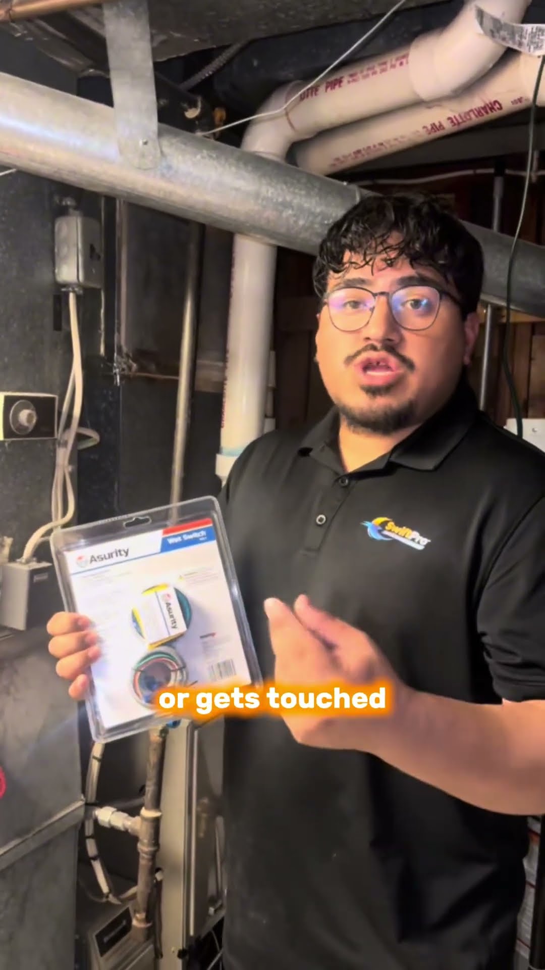 A tech in a black SwiftPro polo and glasses stands in an unfinished basement by the furnace and ductwork, holding up a packaged water-sensing safety switch and explaining how it protects the basement