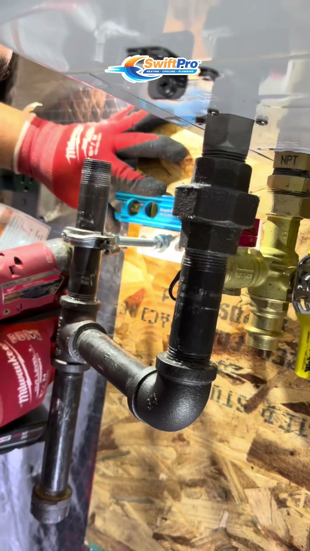 A tankless water heater swap in a basement: a gloved hand levels new black iron gas piping with a union and valves, a tech in a gray cap mounts and connects a wall-hung Navien unit, and a tech on a We