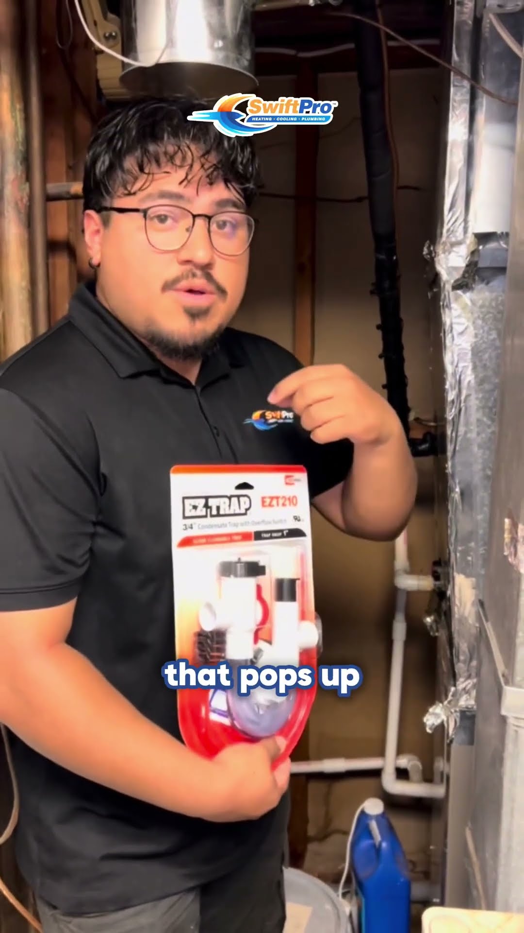 In a basement mechanical room, a tech in a black SwiftPro polo holds up a packaged EZ Trap EZT210 condensate trap with overflow switch and explains it.
