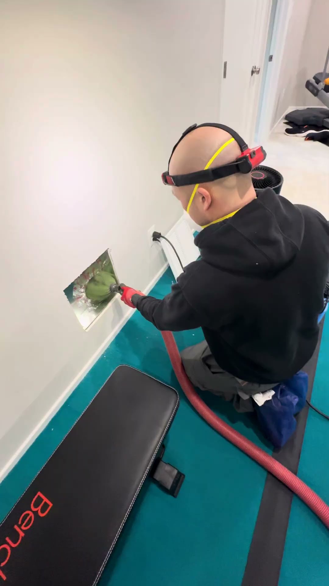 A floor register clogged with dust and debris, then a bald tech with a red headlamp and mask kneels in a home gym running a rotating duct-cleaning brush on a red hose into a wall register.