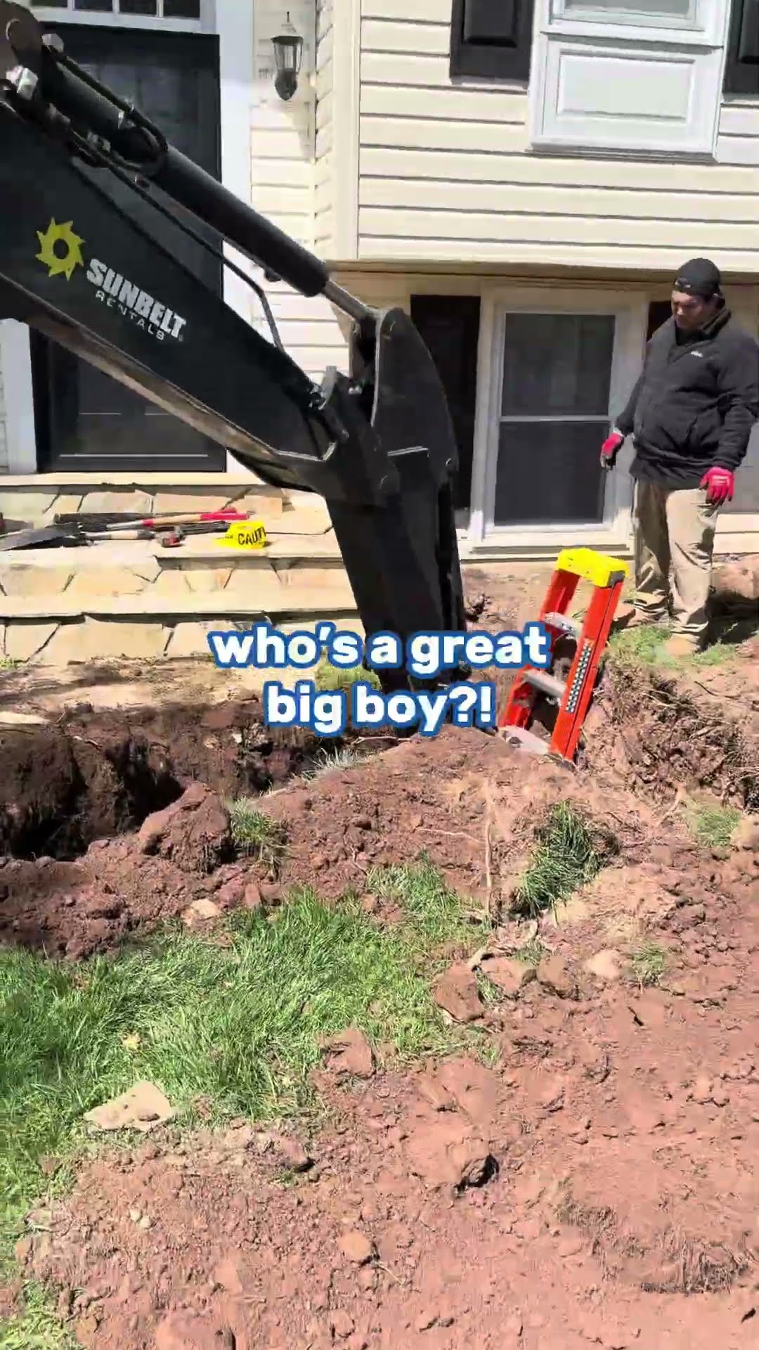 A mini excavator (Sunbelt Rentals branding) digs a trench across a front yard toward a split-level house.