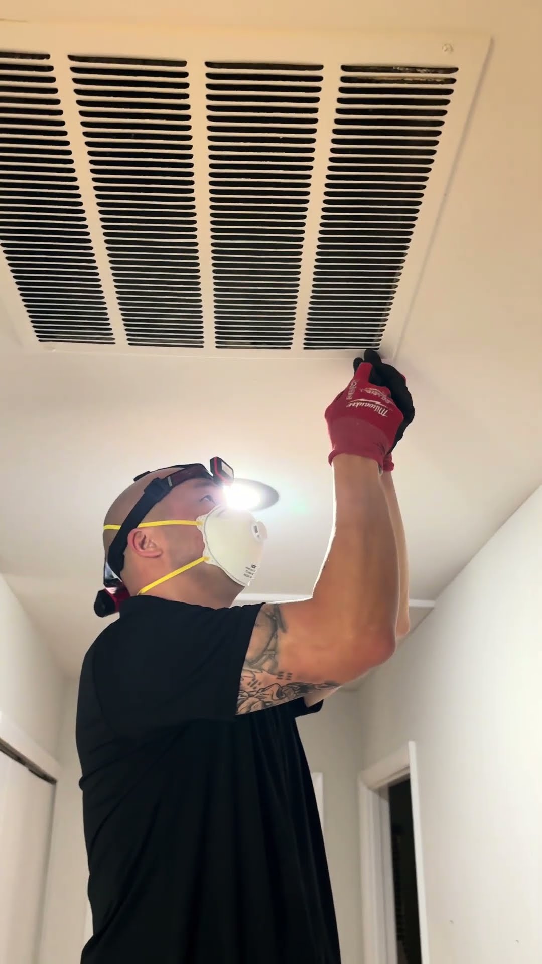 A duct cleaning tech in a headlamp and N95 mask pulls down a ceiling return grille in a hallway.