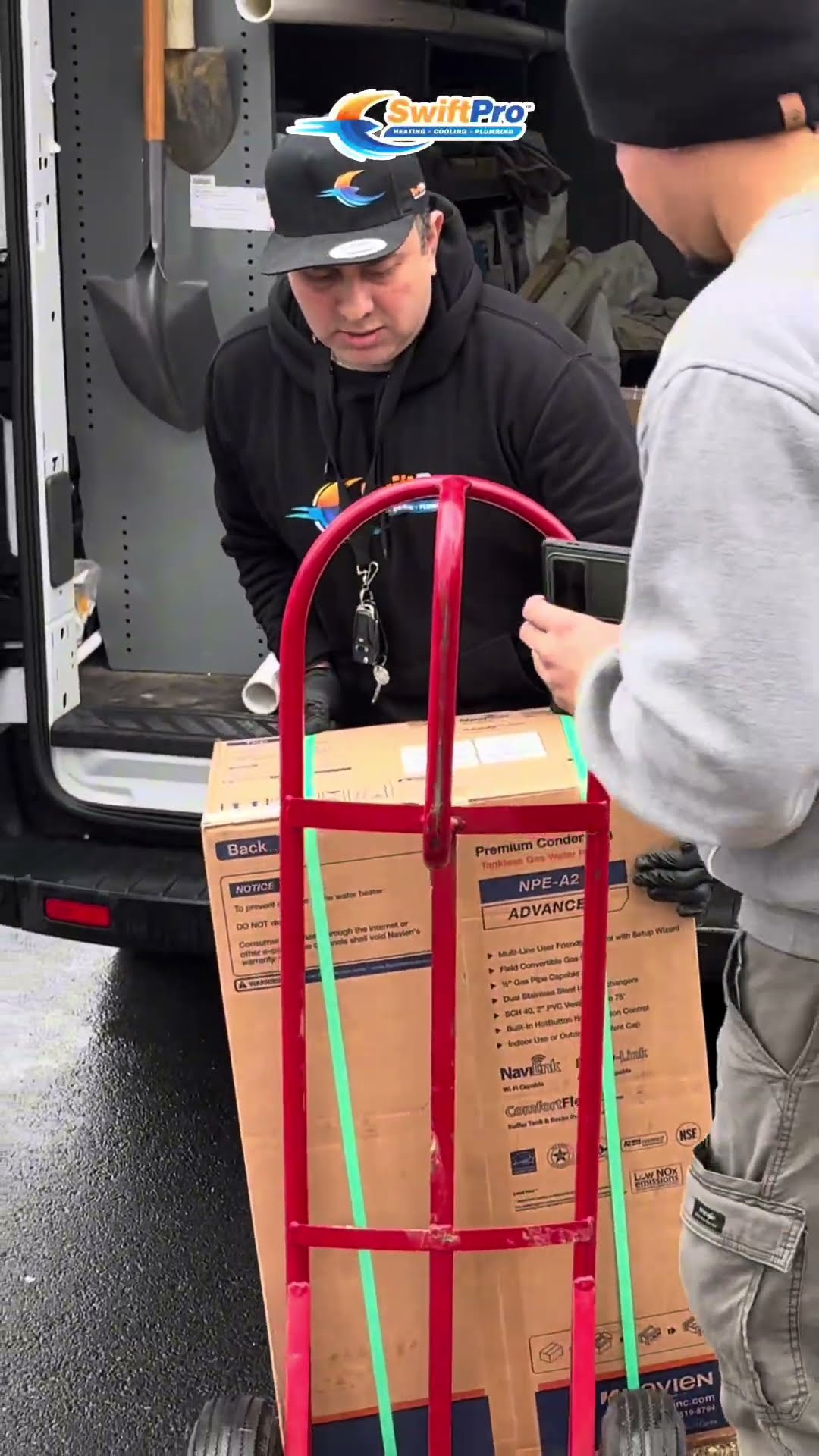 A SwiftPro van parked at a house with a brick pillar; a tech in a SwiftPro hoodie and cap wheels a boxed Navien NPE-A2 tankless water heater off the van on a hand truck; a young tech unboxes parts in
